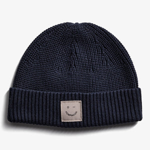 A navy blue knitted beanie lies flat against a white background. The cuff features a sewn patch with a simple face illustration and the text "POMPdeLUX."