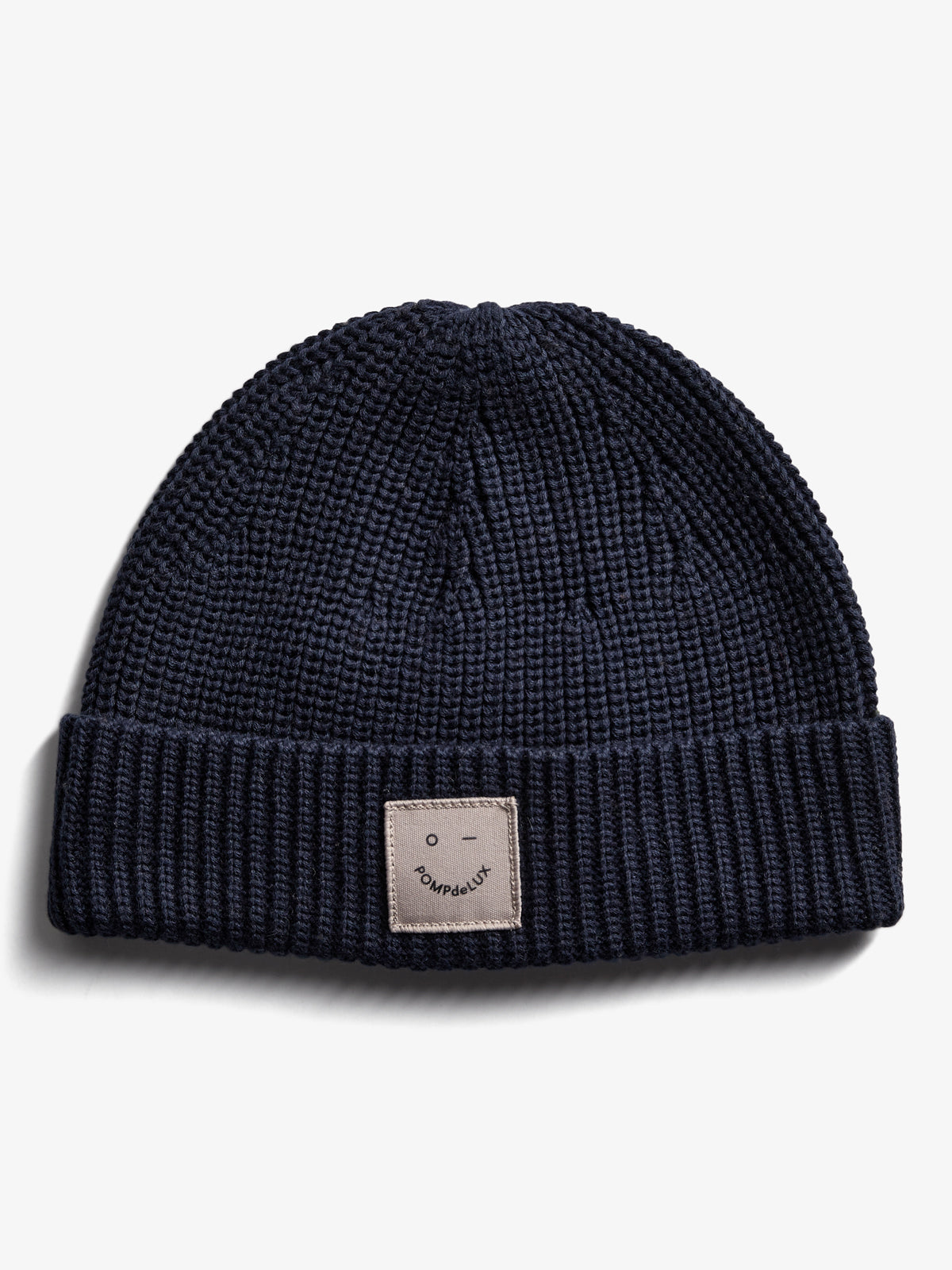 A navy blue knitted beanie lies flat against a white background. The cuff features a sewn patch with a simple face illustration and the text "POMPdeLUX."