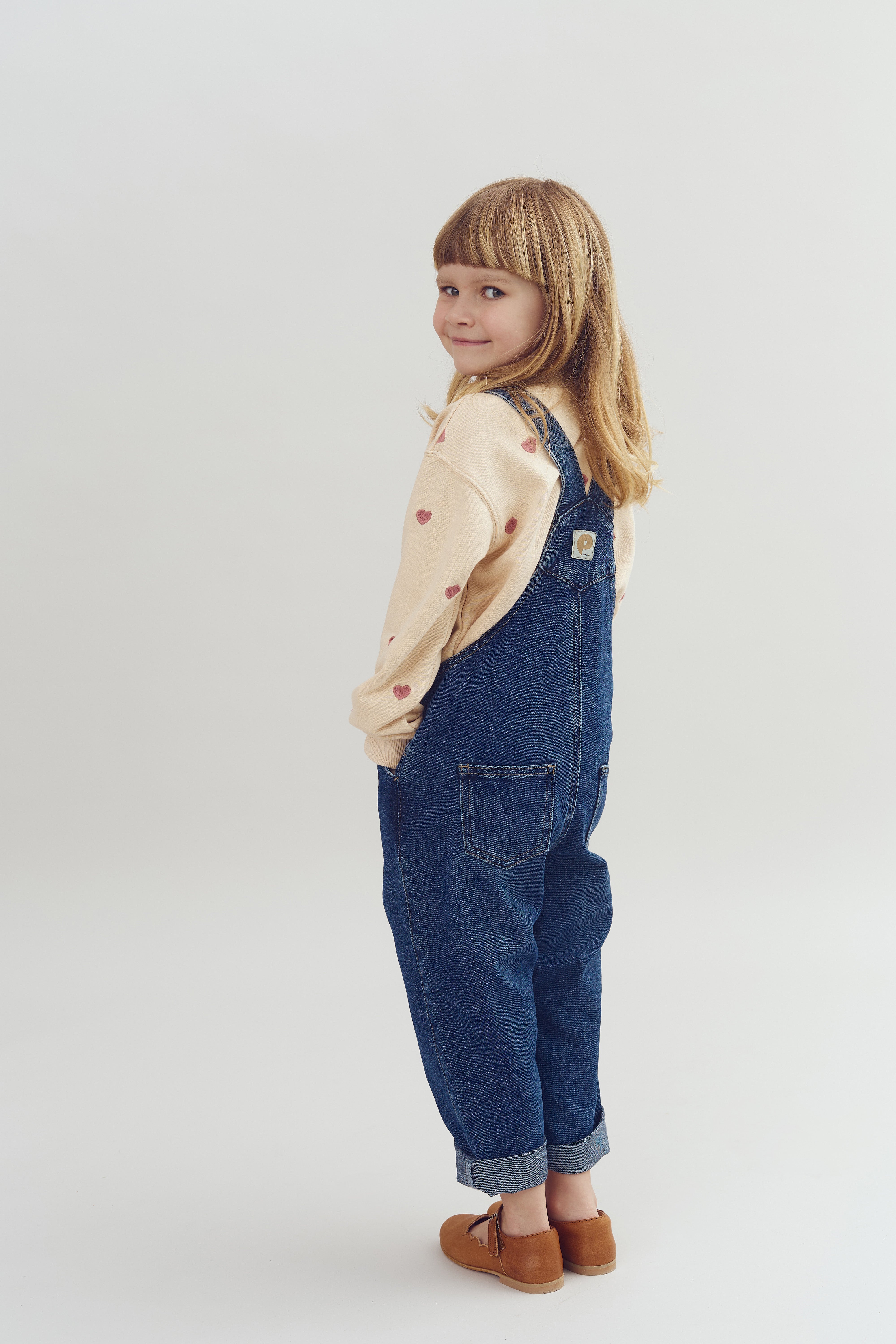 A child in denim overalls and a beige sweater with heart patterns stands sideways, glancing back and smiling, set against a plain white background.