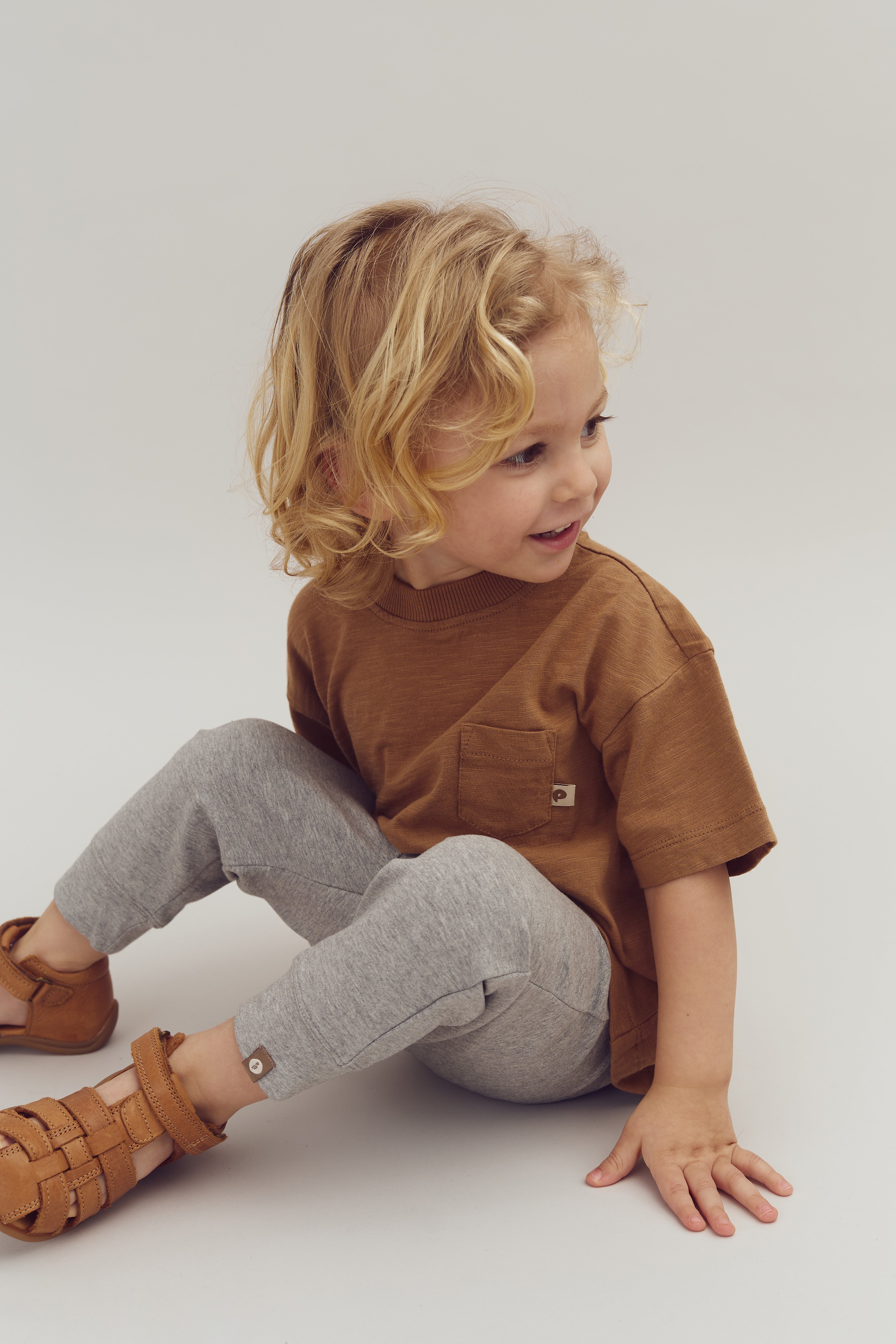 A smiling child sits cross-legged, wearing a brown shirt, gray pants, and brown sandals, on a plain light background. The child gazes to the side, appearing playful and relaxed.