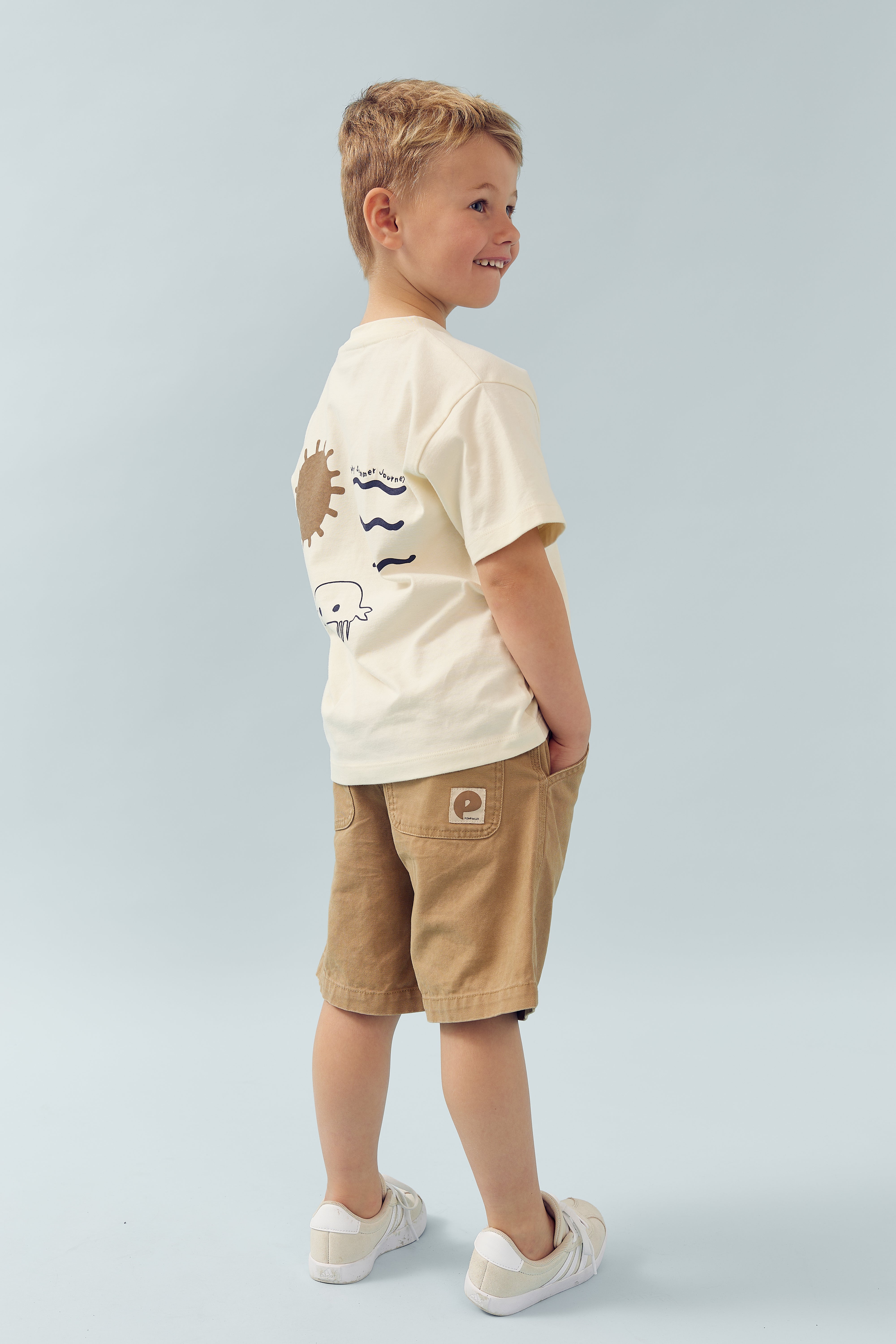 A child wearing a white T-shirt with brown shorts stands smiling, hands in pockets. The shirt has doodles of a whale, waves, and a sun, with text "rugged butts," against a plain light blue background.