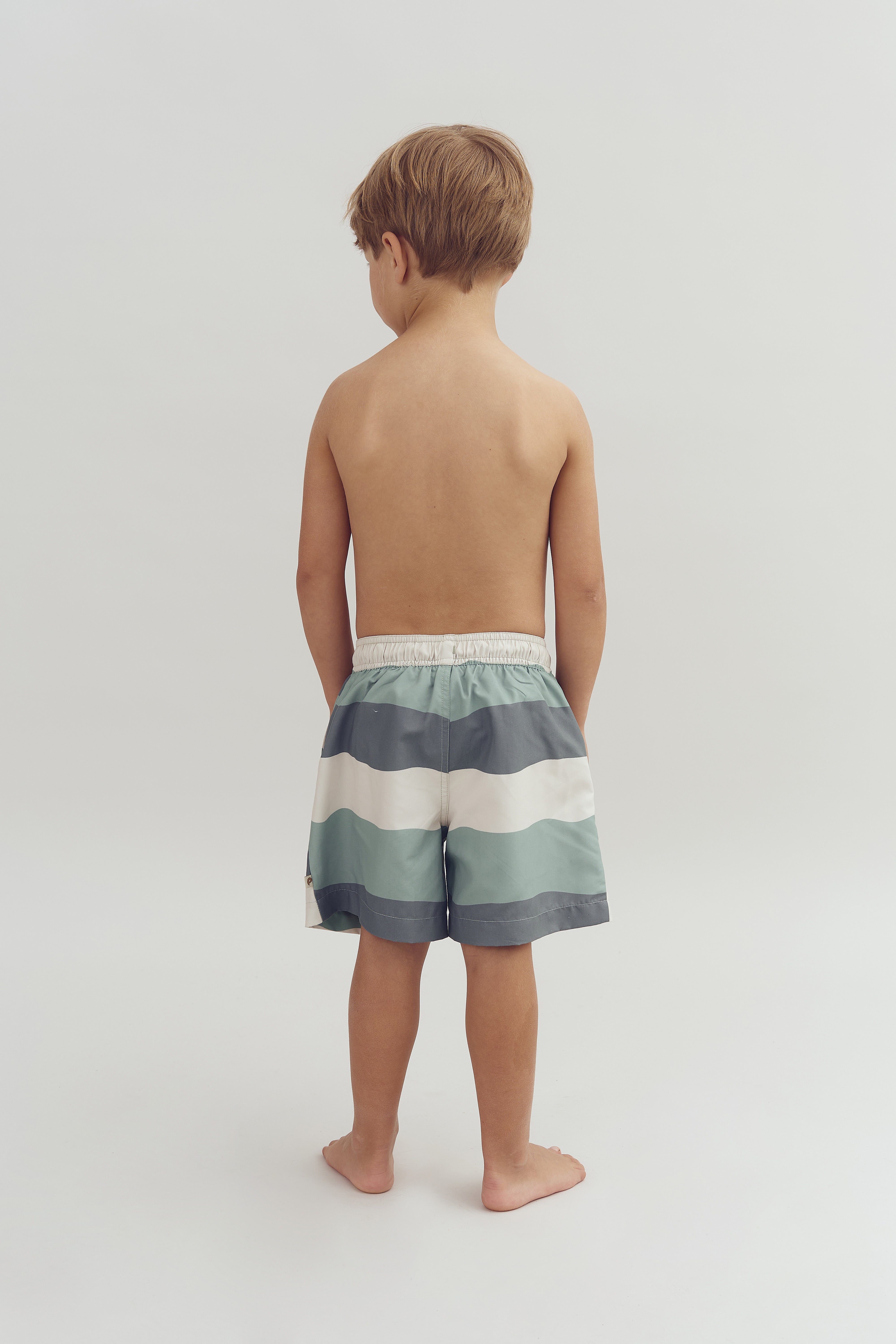 A child stands barefoot, facing away, wearing striped shorts in a minimal, light-colored environment.