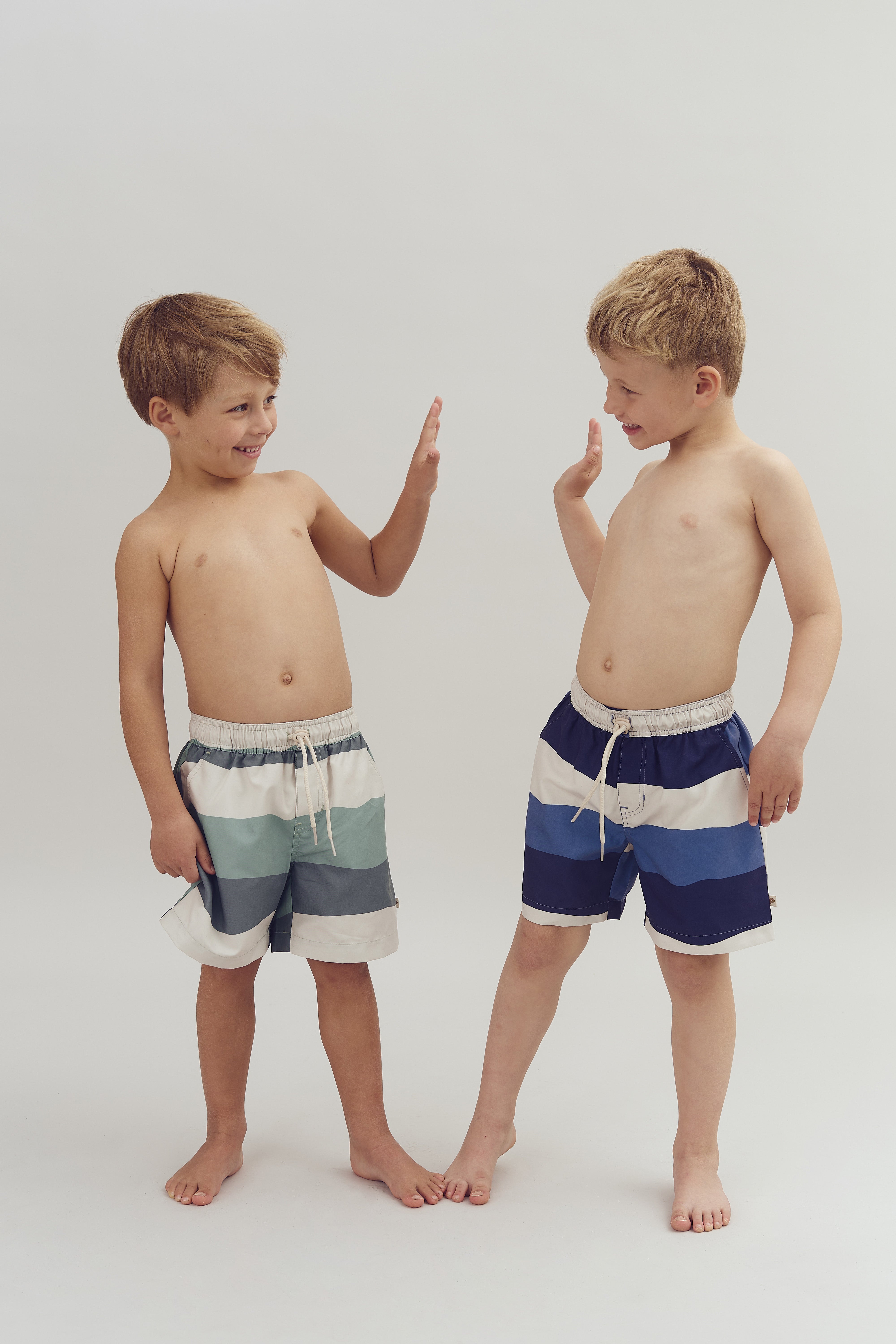 Two boys, wearing striped swim shorts, are facing each other and exchanging high-fives. They are standing barefoot on a plain, light background.