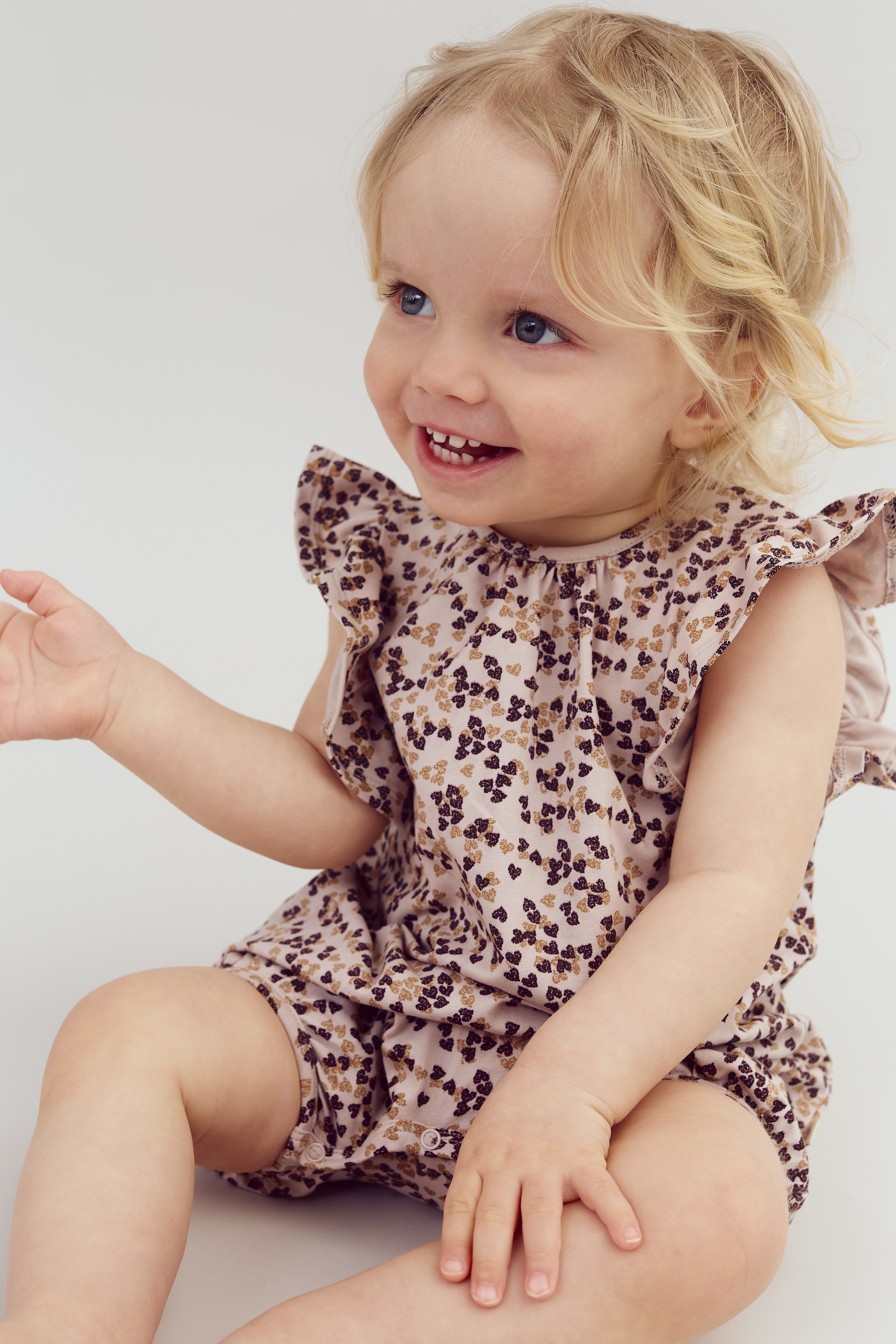 A young child with light hair smiles happily, sitting and wearing a floral-patterned outfit with ruffled sleeves, on a plain white background.