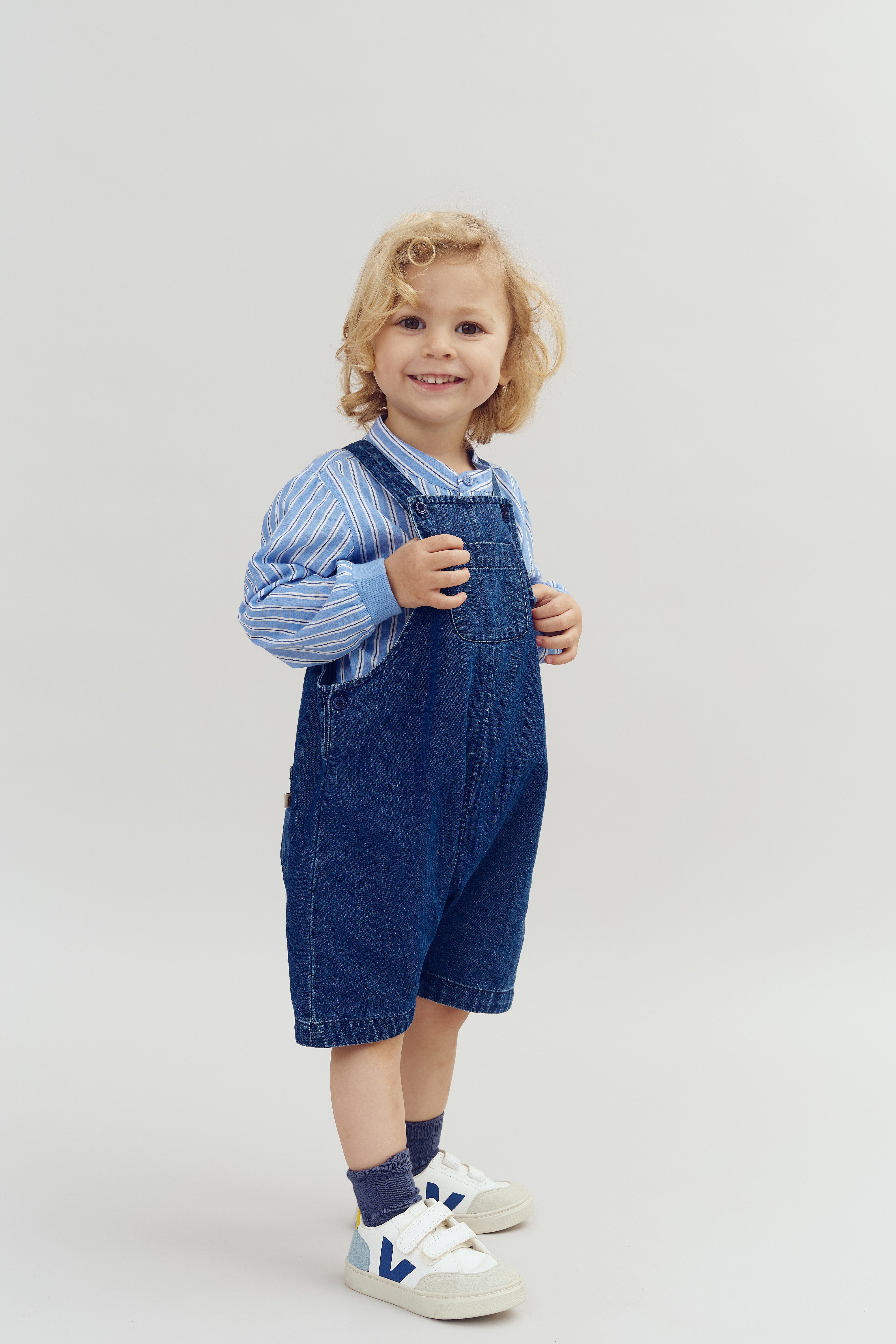 A young child with curly blonde hair smiles while standing, wearing a blue striped shirt, denim overalls, and white sneakers, set against a plain light gray background.