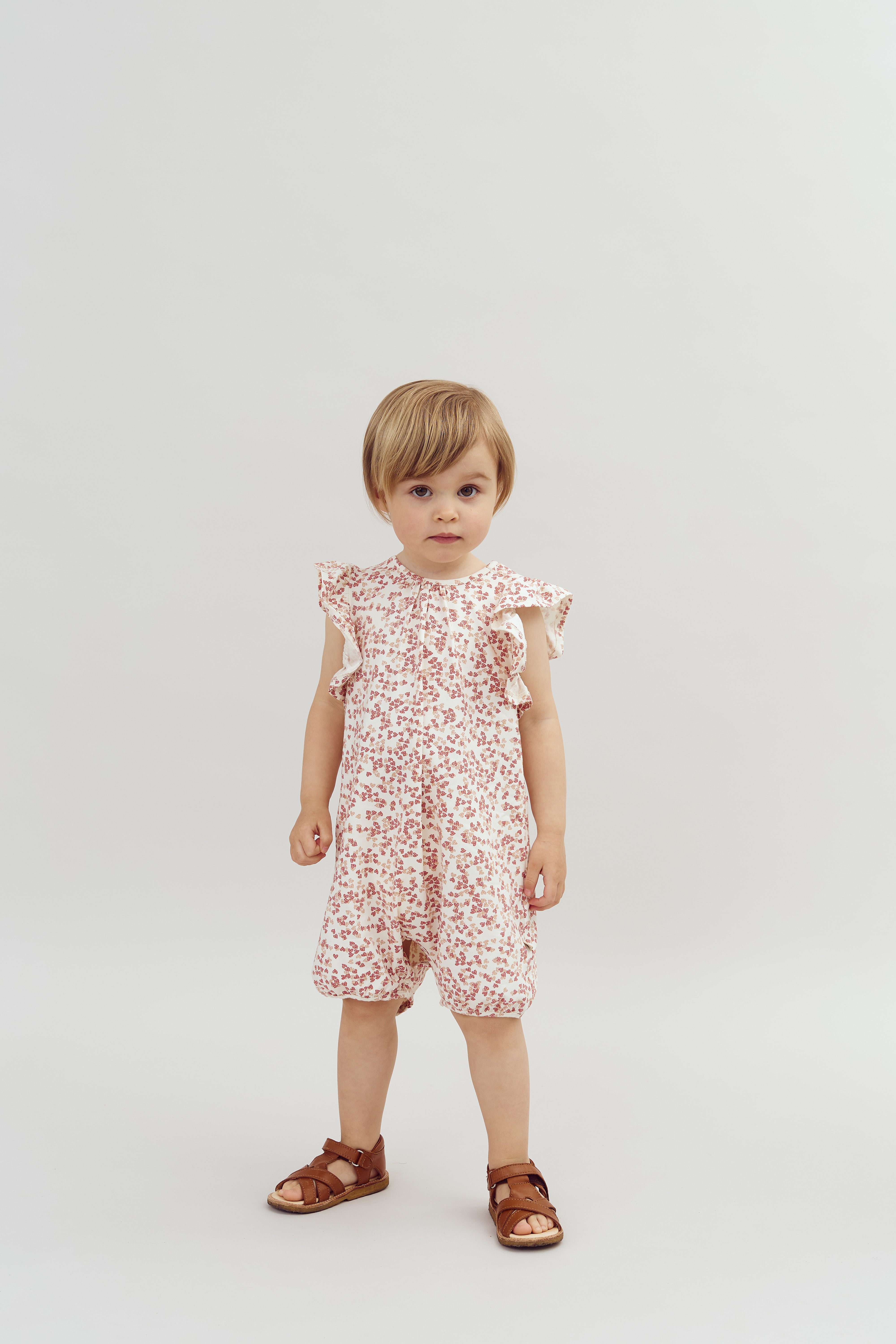 A young child stands in a light-colored studio, wearing a white floral romper and brown sandals. The background is plain and minimalistic, emphasizing the child's outfit.