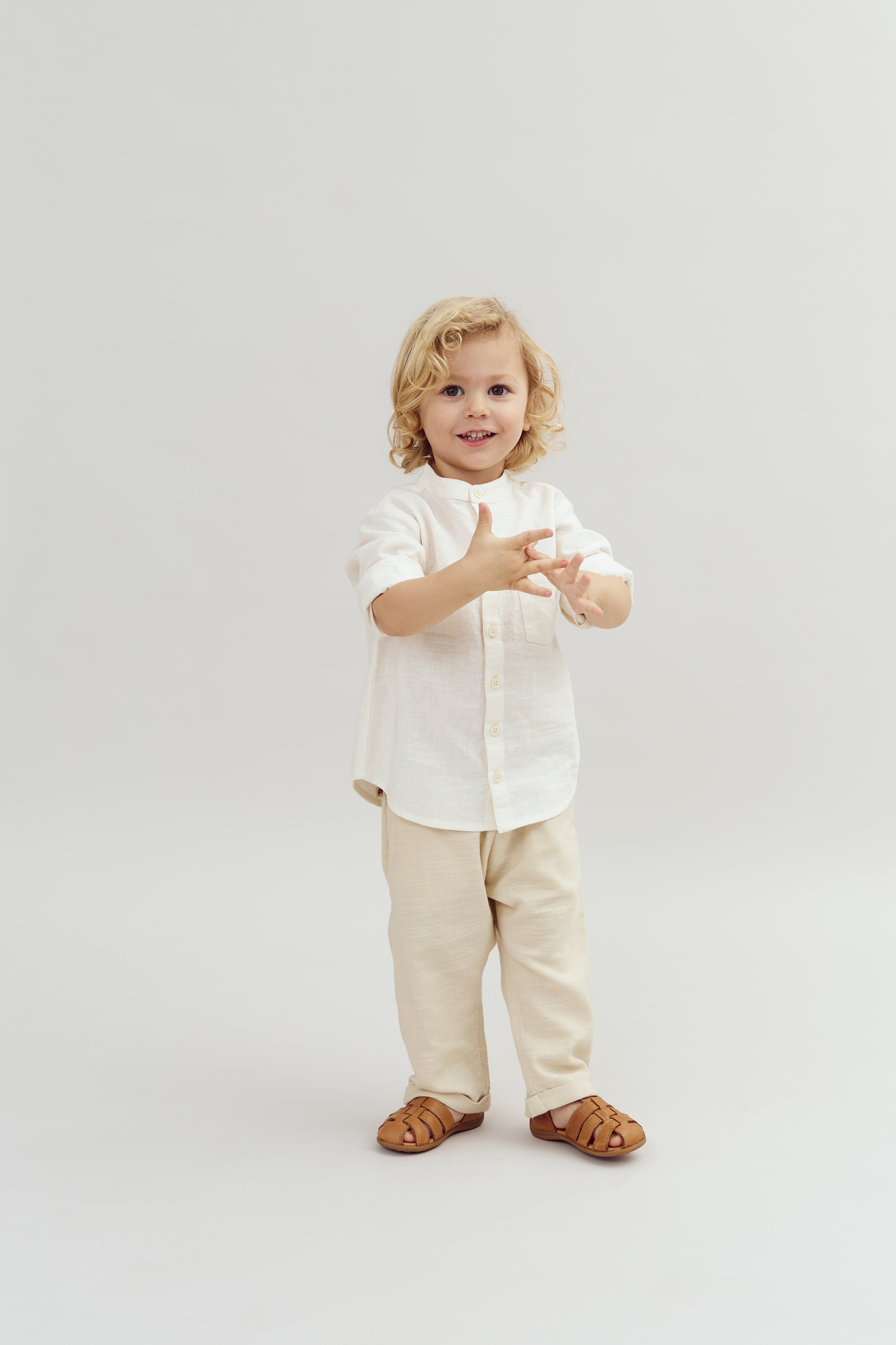 A child with blonde hair is smiling and gesturing with hands, wearing a light-colored outfit and brown sandals, standing against a plain white backdrop.