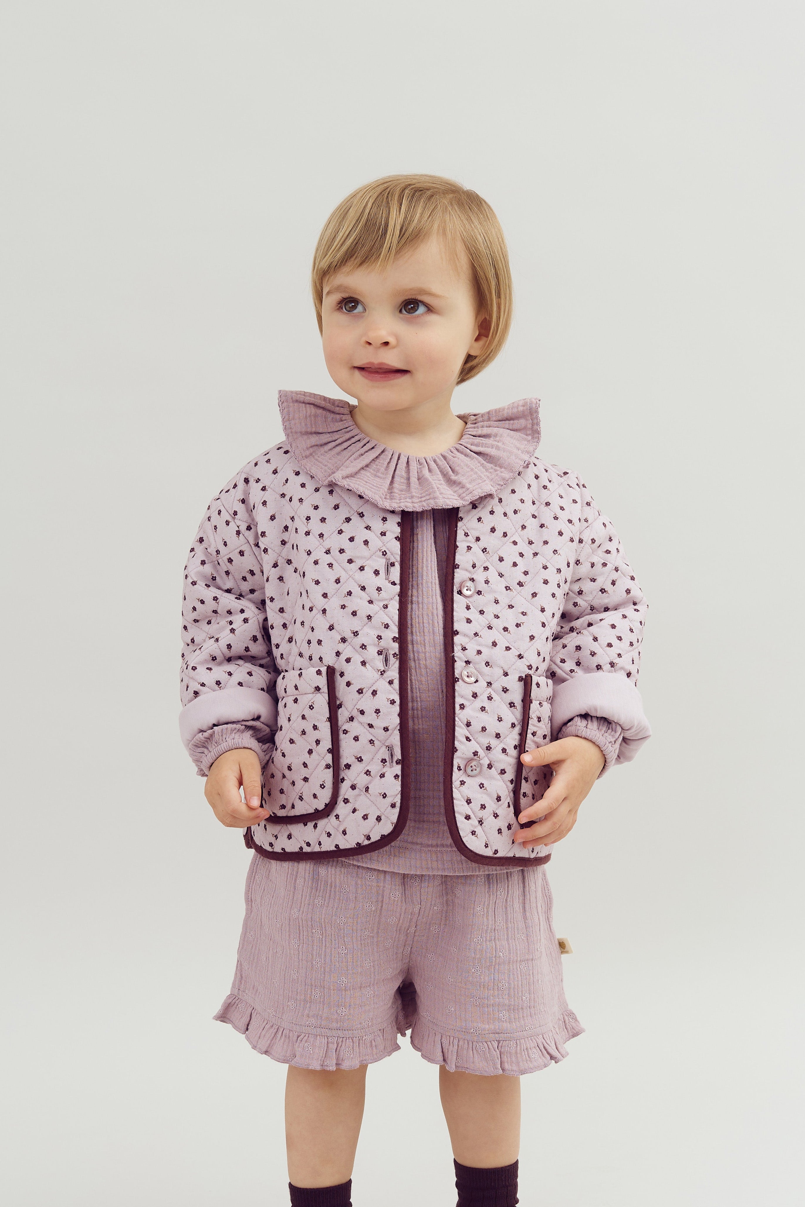 A young child is standing, wearing a lavender quilted jacket with small floral patterns and matching ruffled shorts. The child looks slightly to the side, set against a plain background.