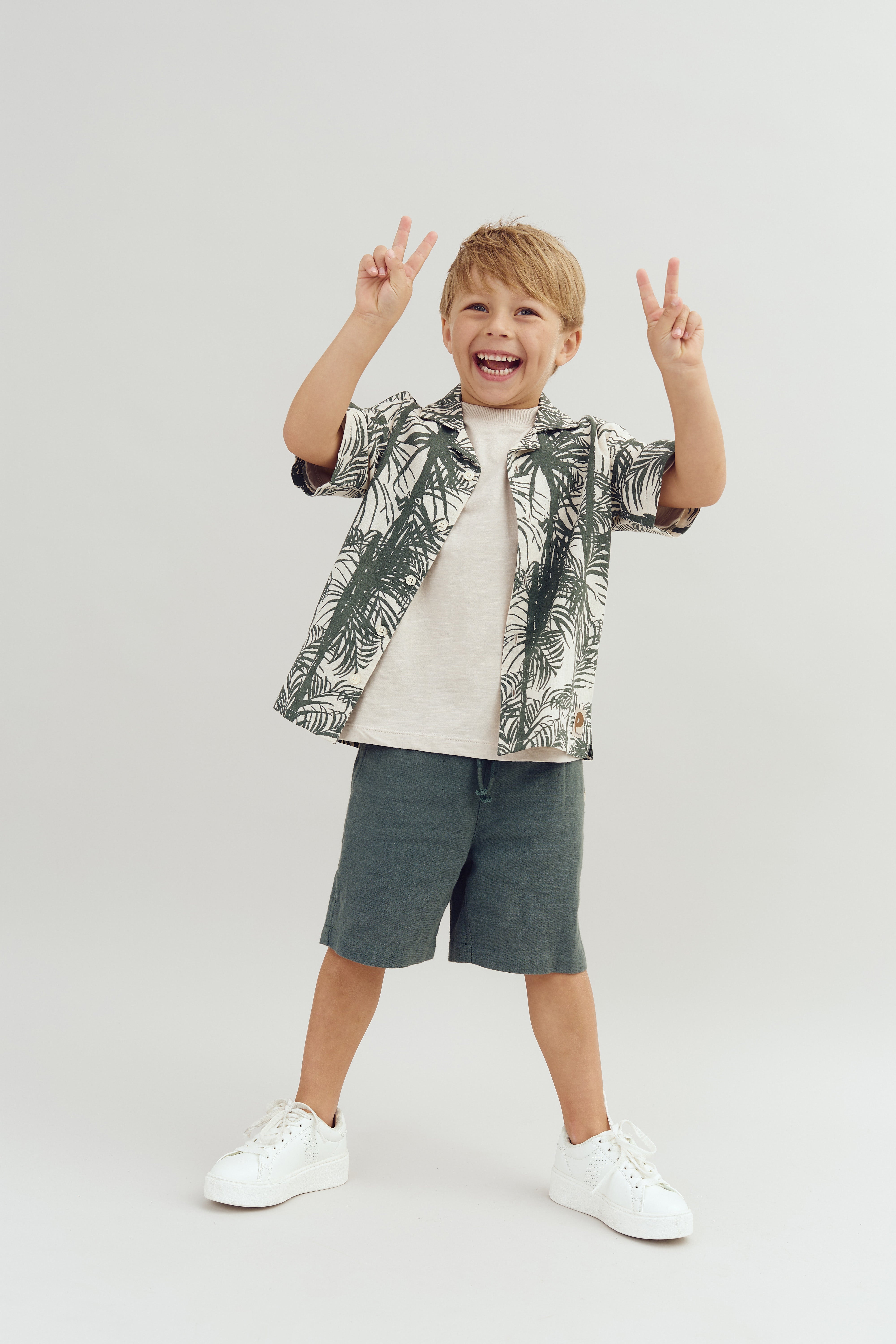 A smiling child wearing a patterned shirt and shorts makes peace signs with both hands in front of a plain, light background.