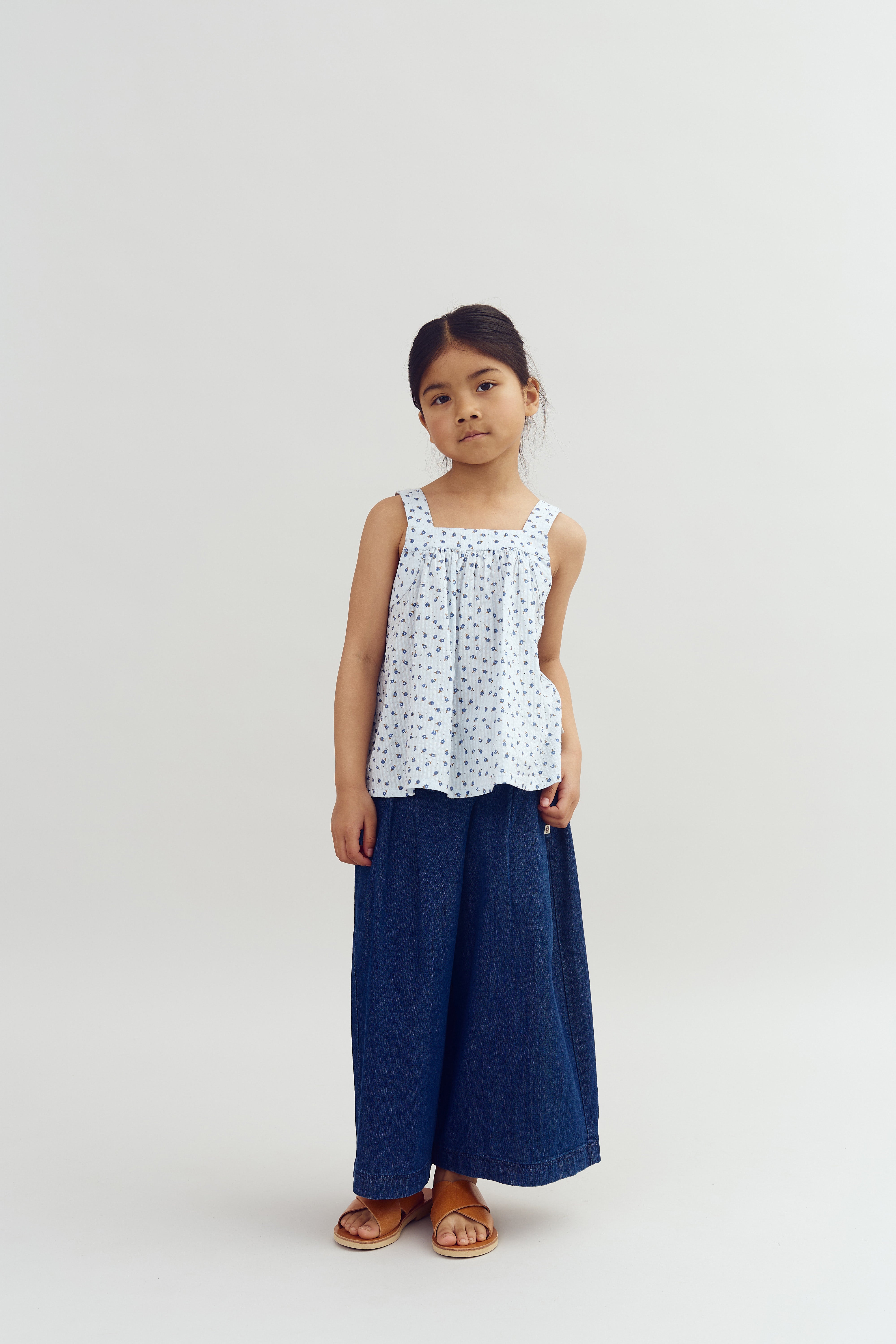 A child stands, wearing a white patterned top, wide blue jeans, and brown sandals, against a plain white background, looking directly at the viewer.