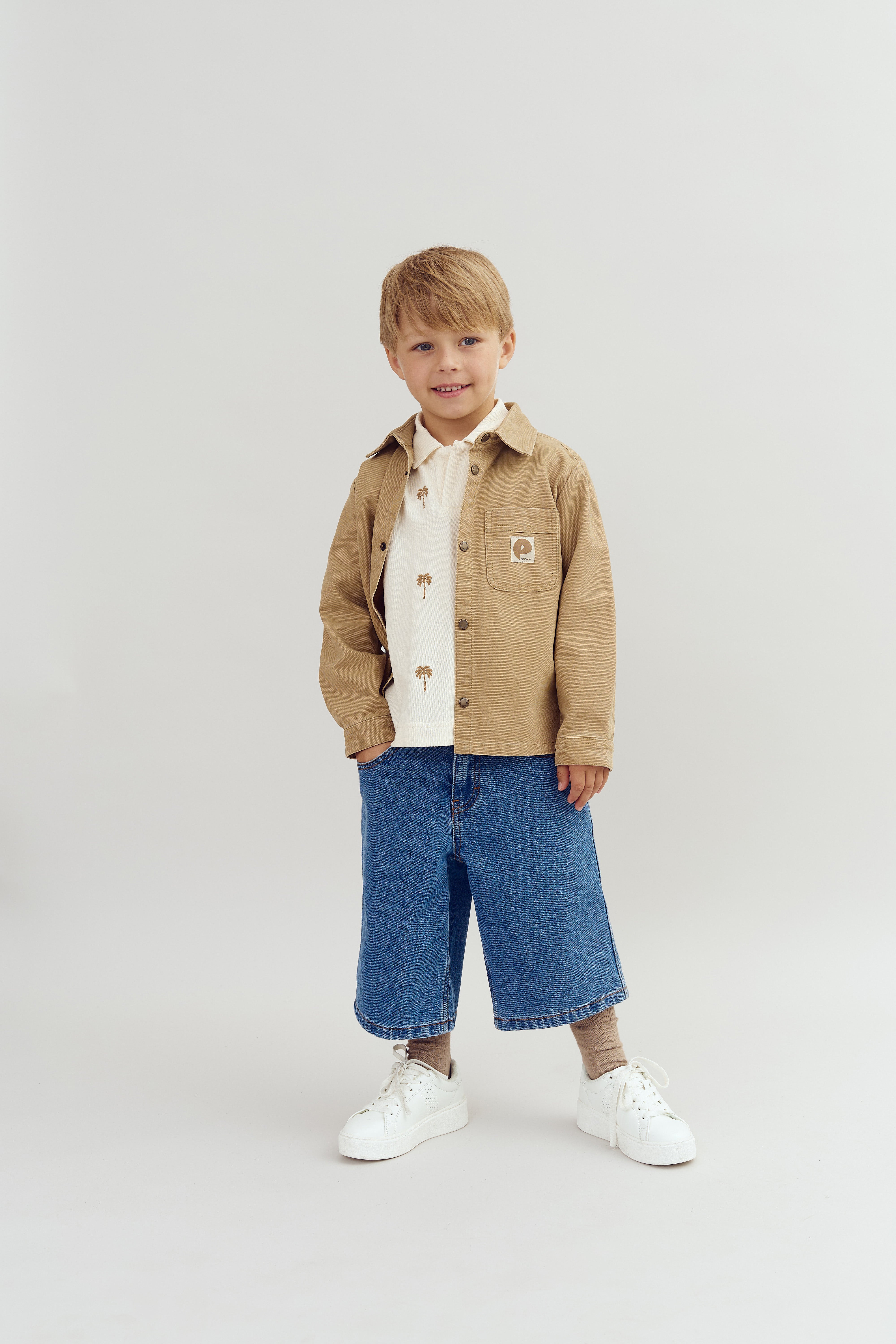 A young child stands smiling, wearing a tan jacket, palm tree shirt, wide blue jeans, and white sneakers, against a plain light background.