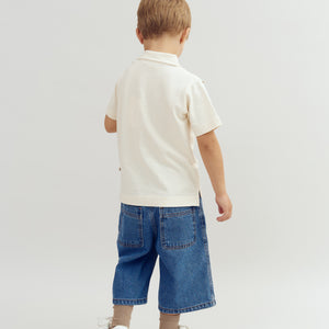 A child stands facing away, wearing a white shirt, denim shorts, and white shoes, in a minimalist, light-colored studio setting.