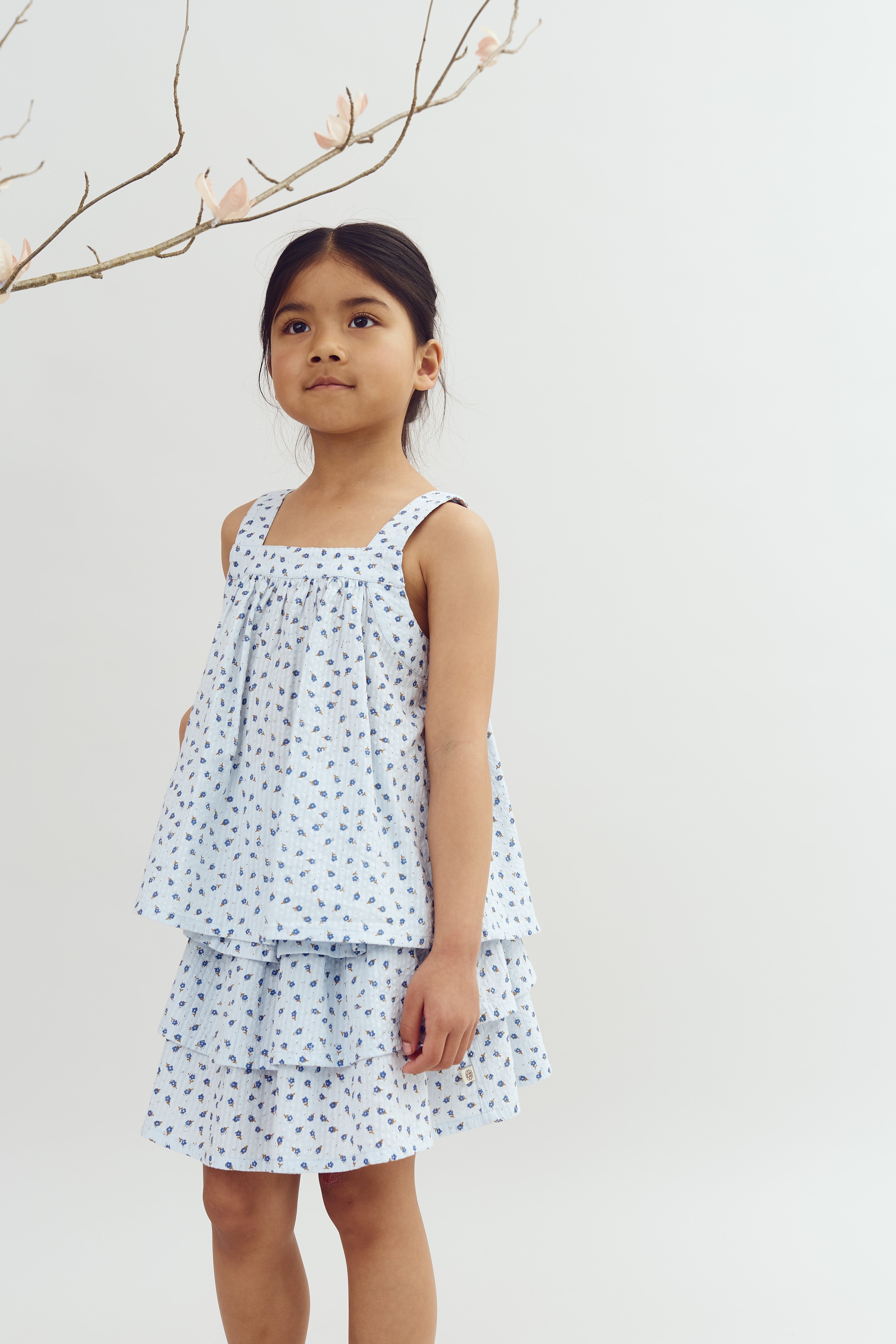 A young girl stands looking upward, wearing a light blue, floral-patterned dress. She is beneath a branch with sparse pink blossoms, set against a plain white background.