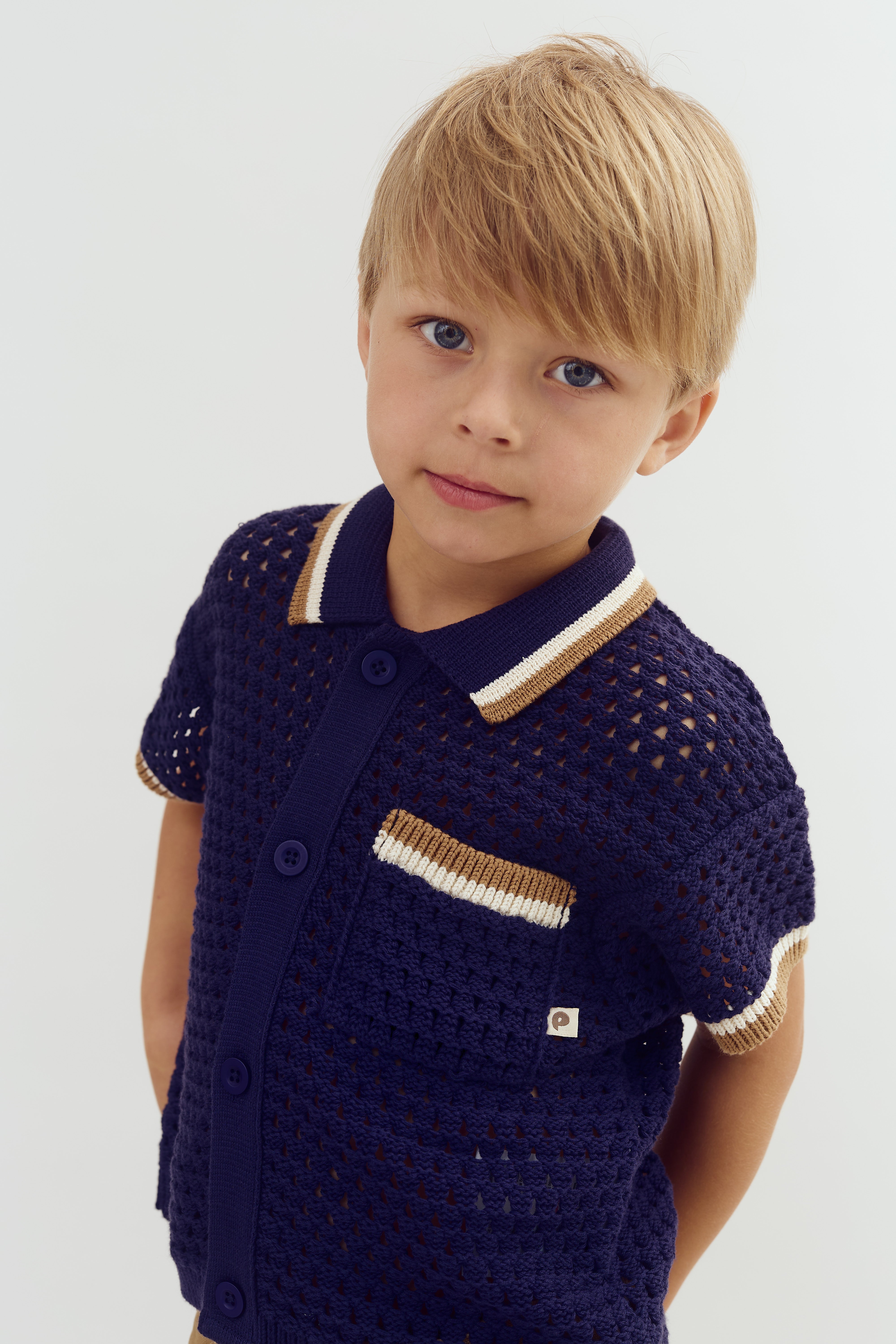 A child wearing a navy, short-sleeved crochet shirt with a collar and pocket, featuring beige and white trim, gazes forward against a plain background.