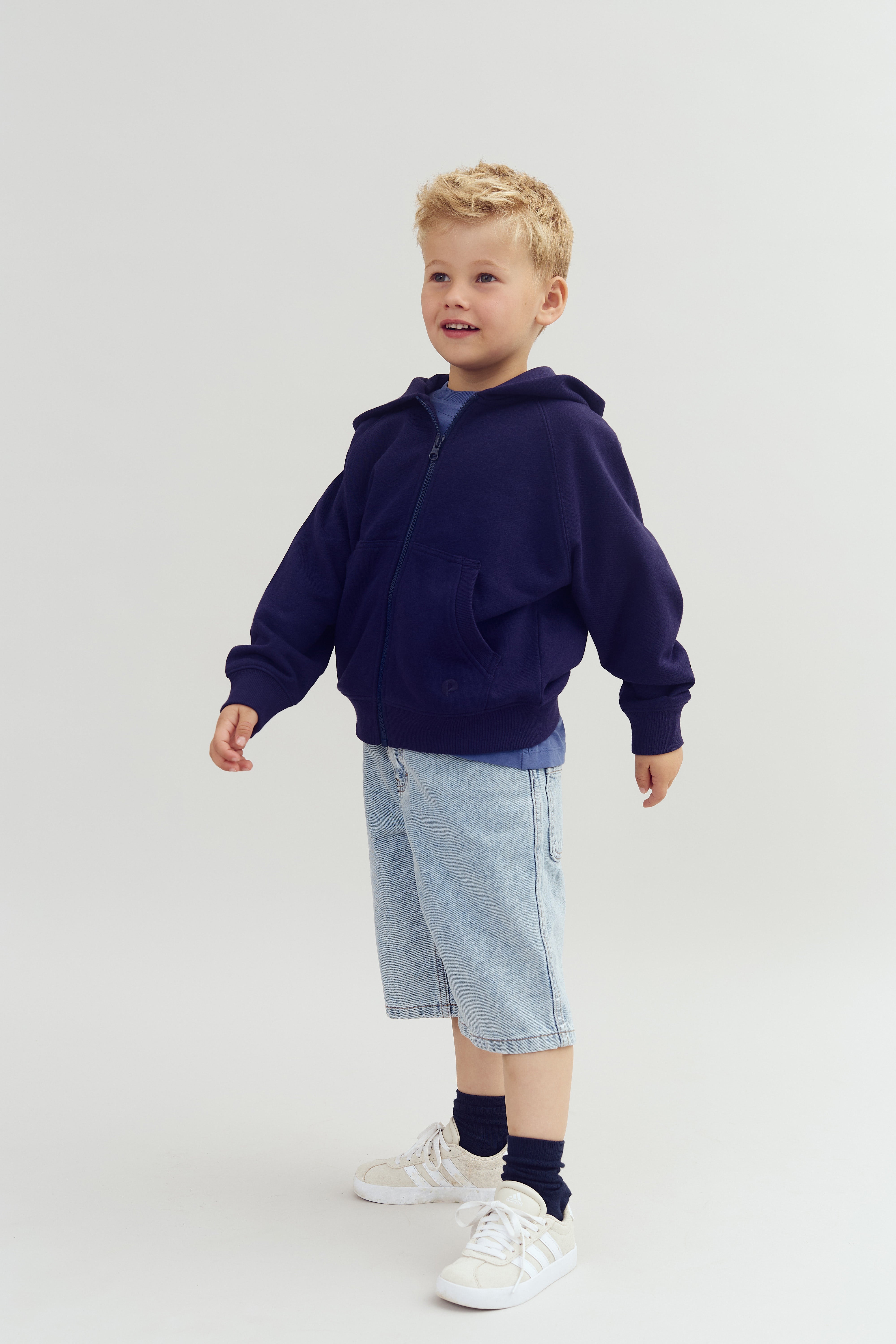 Child in a dark blue hoodie and light denim shorts stands smiling in a studio setting. The child wears white sneakers and dark socks, with a minimalistic light background.