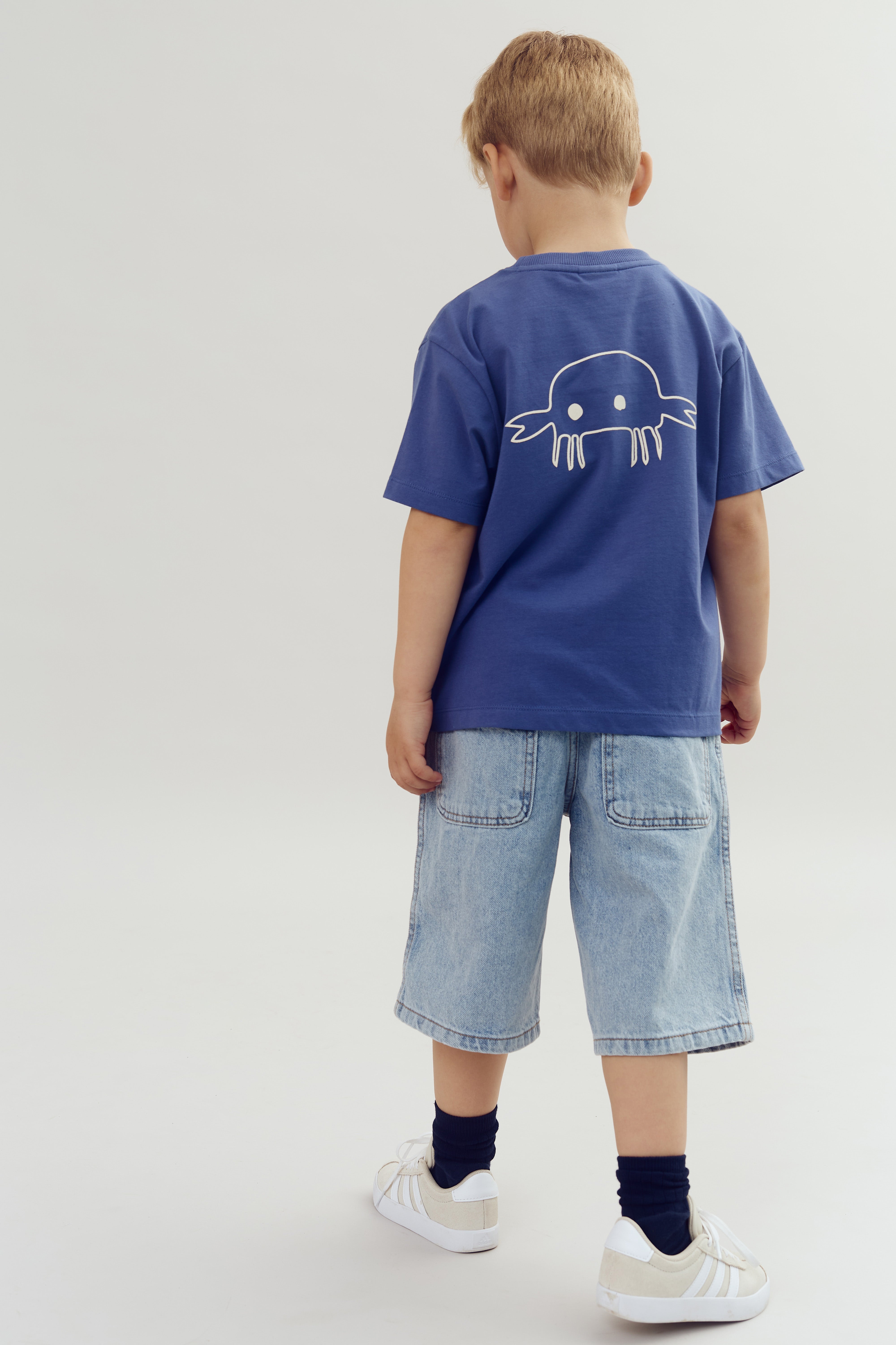 Child facing away, wearing a blue shirt with a simple white crab design on the back, light denim shorts, and white sneakers, standing in a plain, light-colored room.