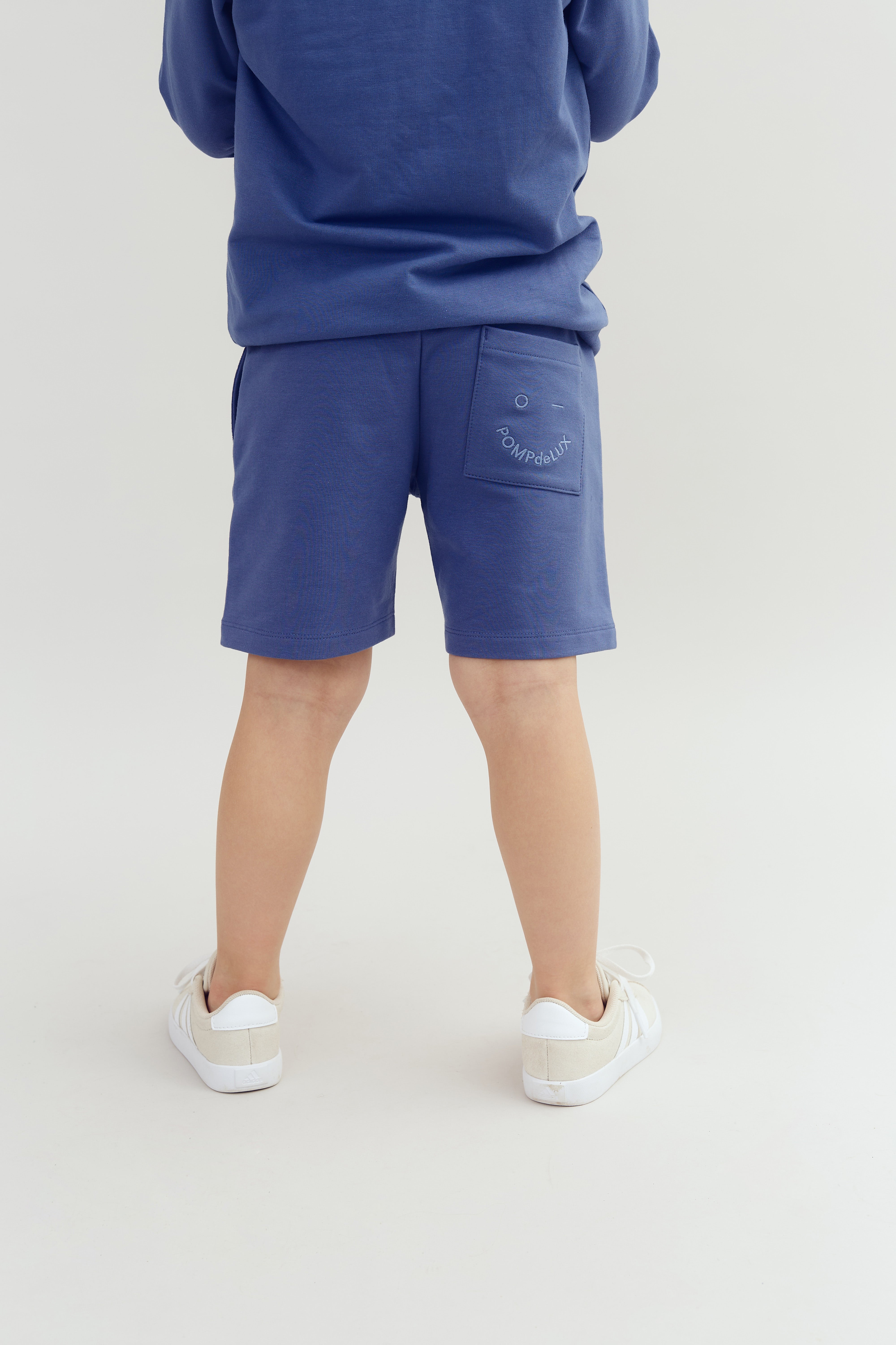 A child stands in blue shorts with "POMPdeLUX" on a pocket, wearing a matching blue top and white sneakers, against a neutral backdrop.
