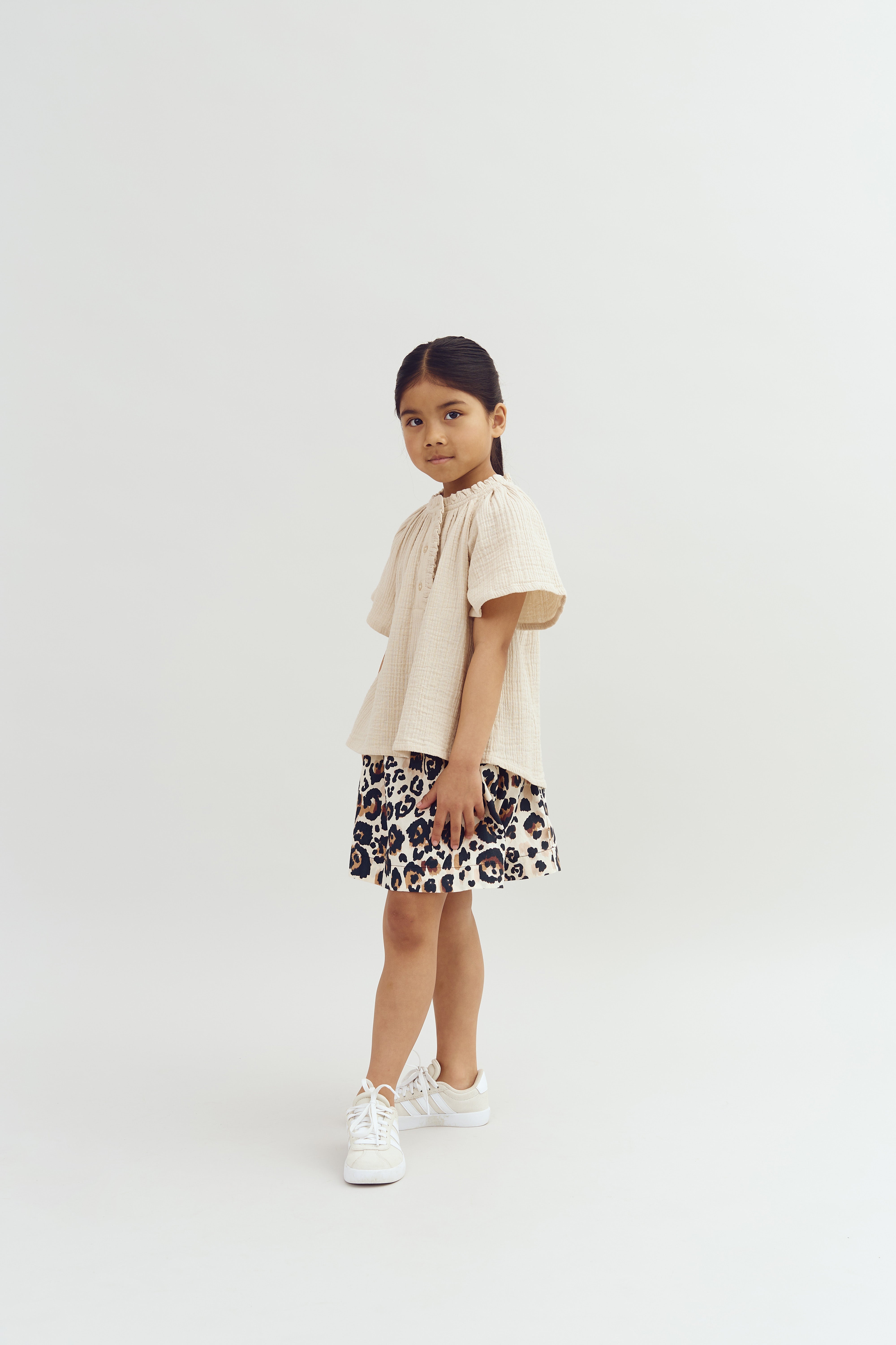 A young girl stands confidently, wearing a beige top and leopard-print skirt with white sneakers, set against a plain light background.