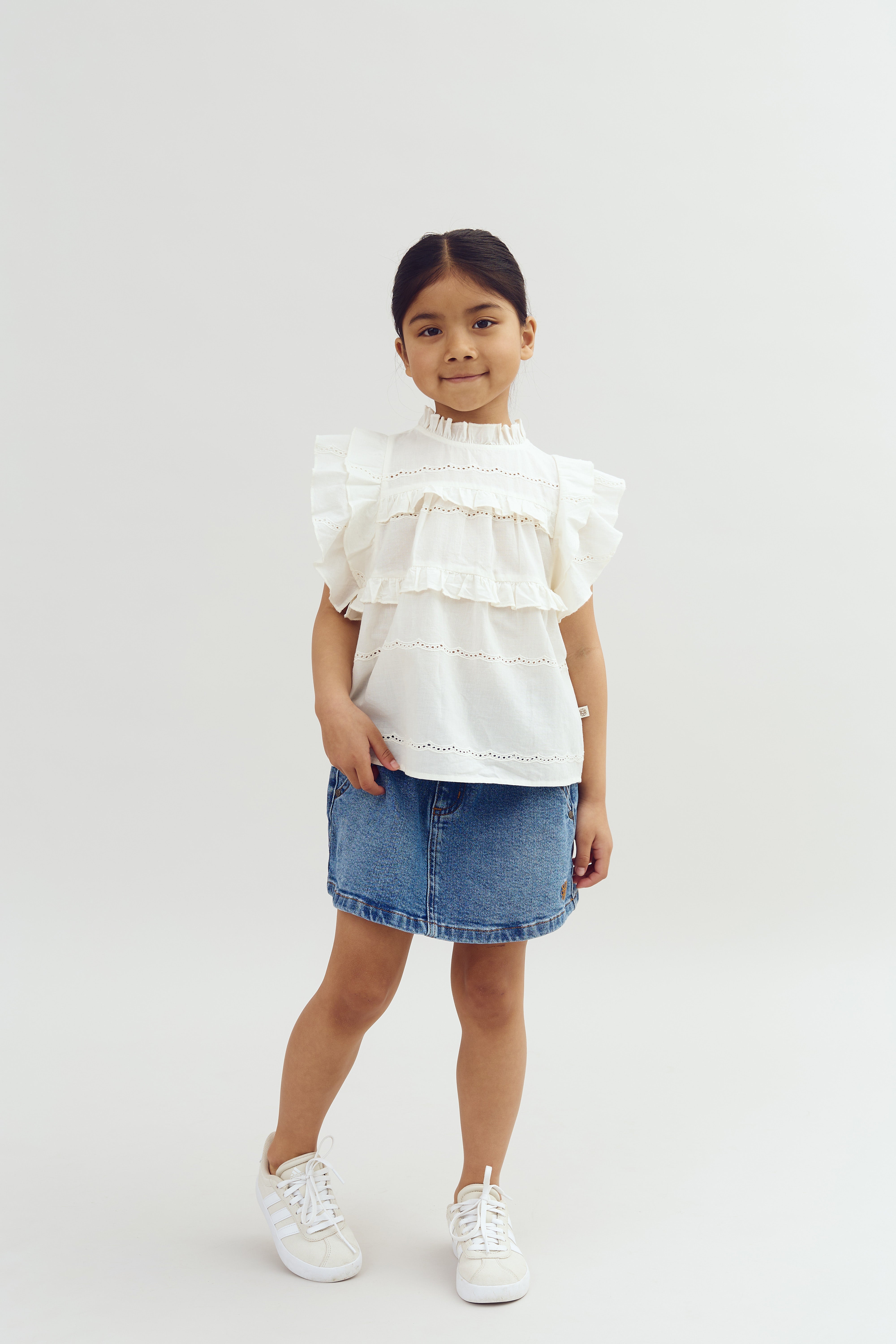 A young girl stands confidently, wearing a white blouse with ruffled layers and a denim skirt. She is against a simple, light-colored background, exuding a cheerful demeanor.