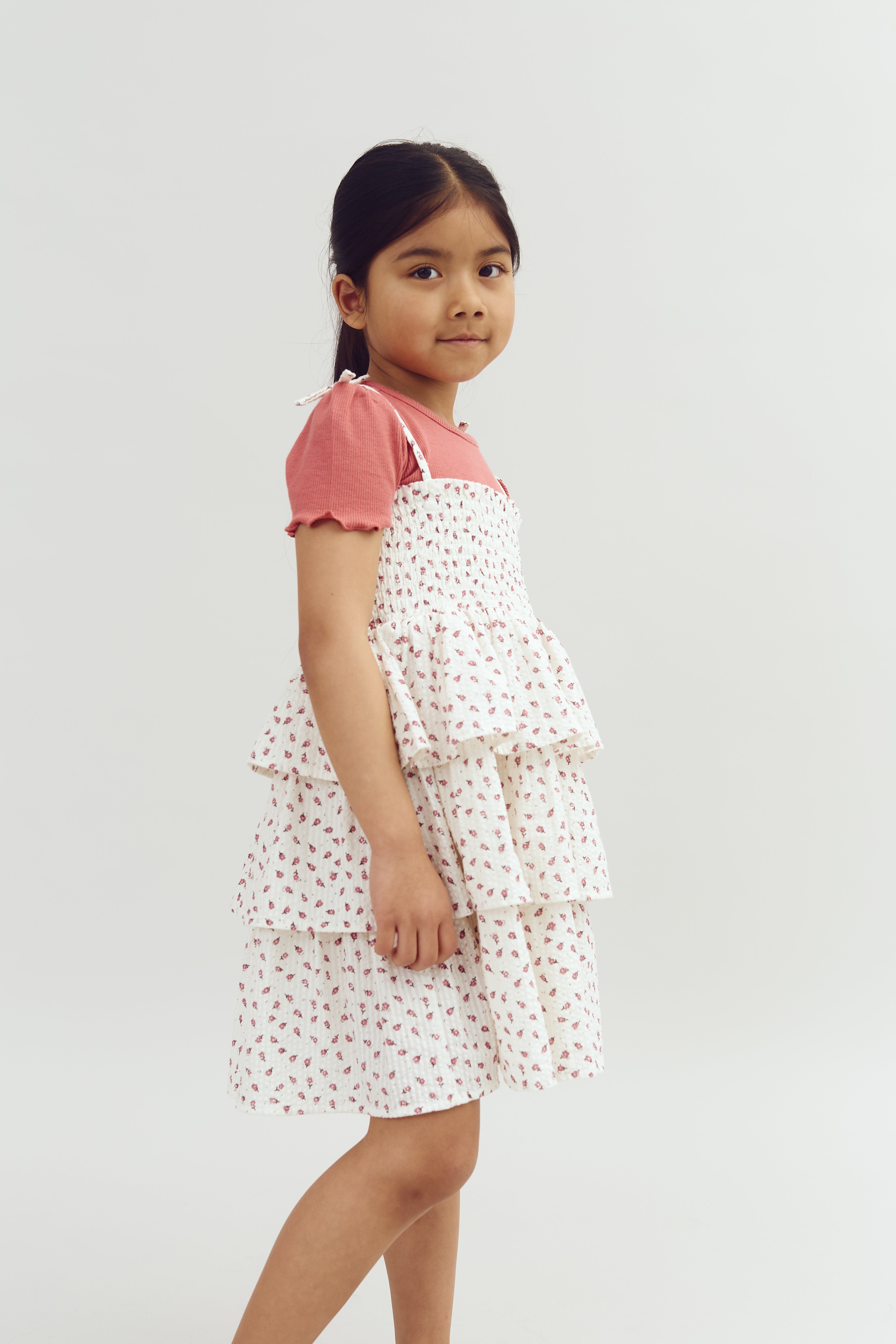 A young girl stands in a white dress with floral patterns over a pink shirt, set against a plain white background.