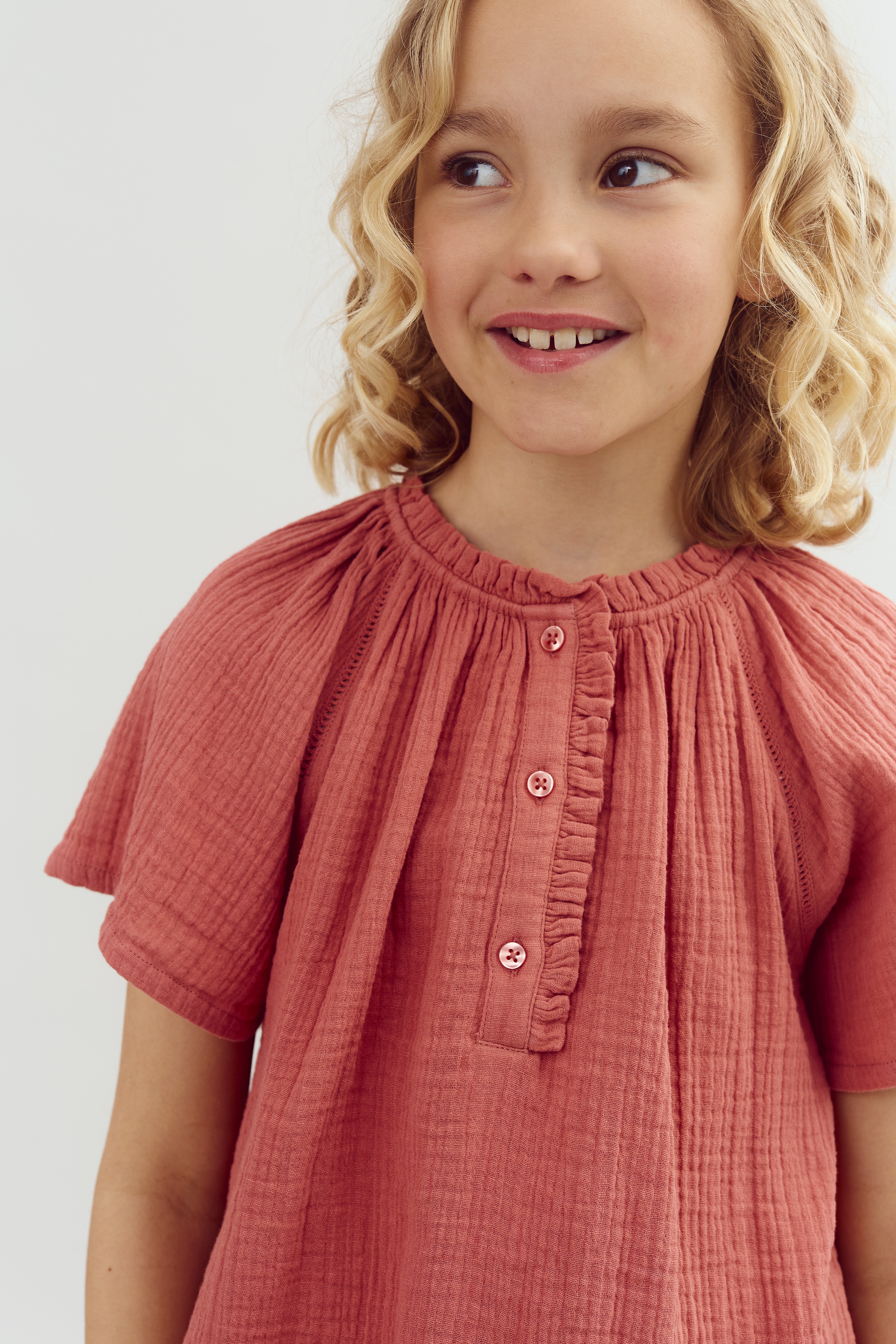 Child wearing a textured, pink button-up shirt smiles slightly while looking to the side, showcasing short, curly blonde hair, against a plain, light-colored background.