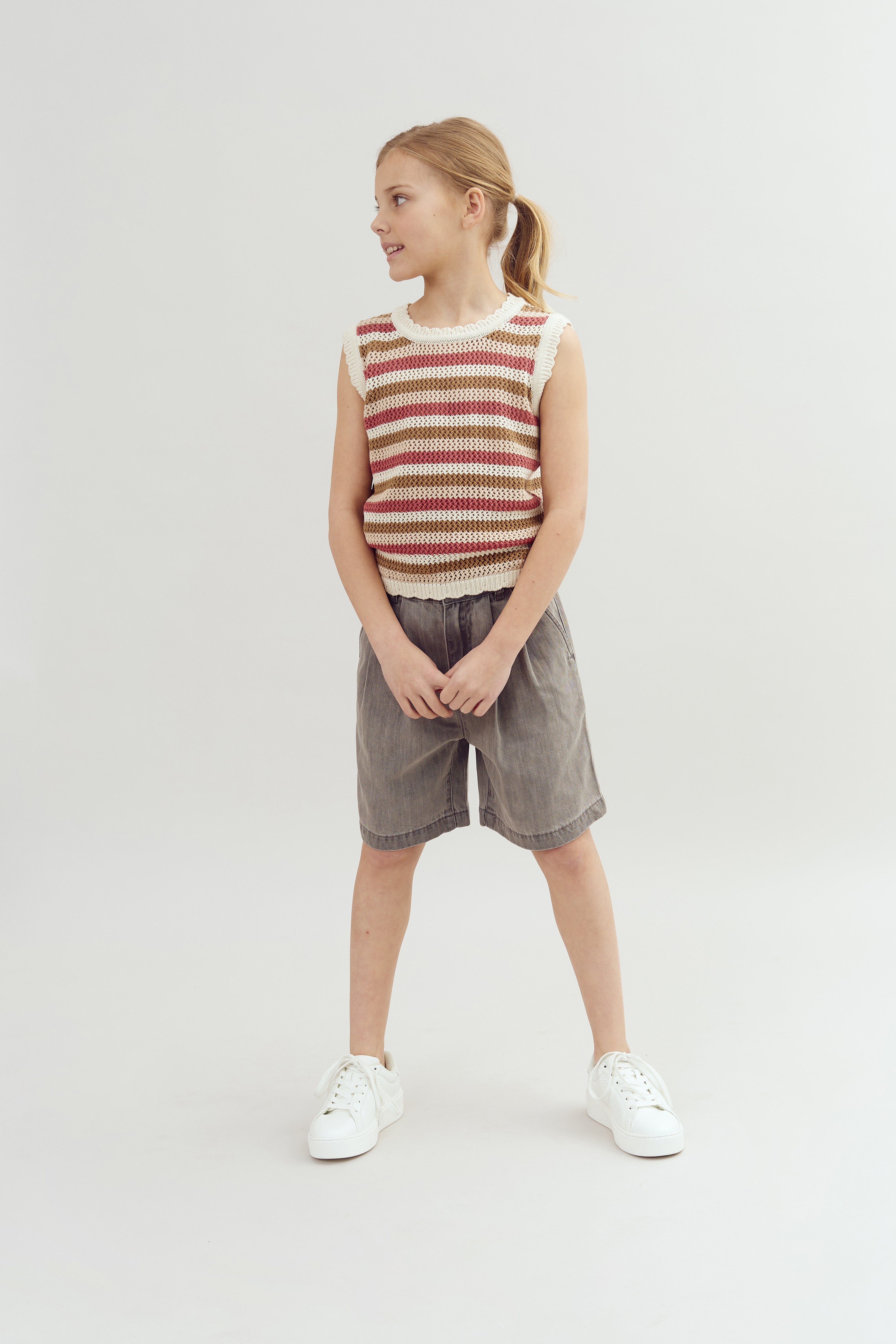 A child stands smiling, wearing a striped sleeveless top, gray shorts, and white sneakers, against a plain gray background.